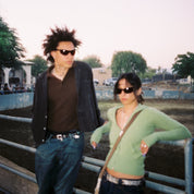 Two people standing behind a metal fence with a blurred background. Heat Polarized Bronze sunglasses