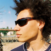 Person wearing sunglasses and a blue shirt with a cross necklace, standing outdoors.
