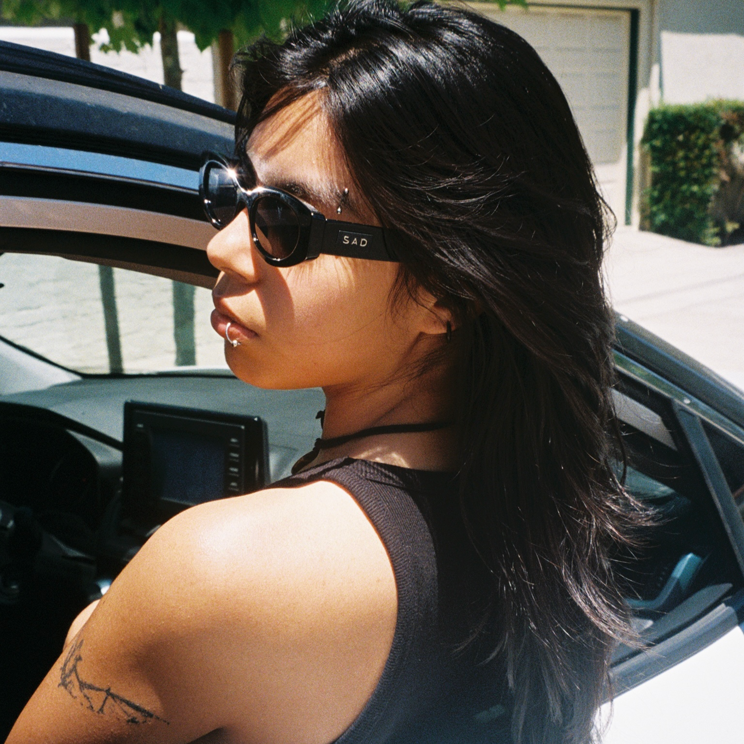 Person sitting in a car wearing sunglasses with a house and tree in the background