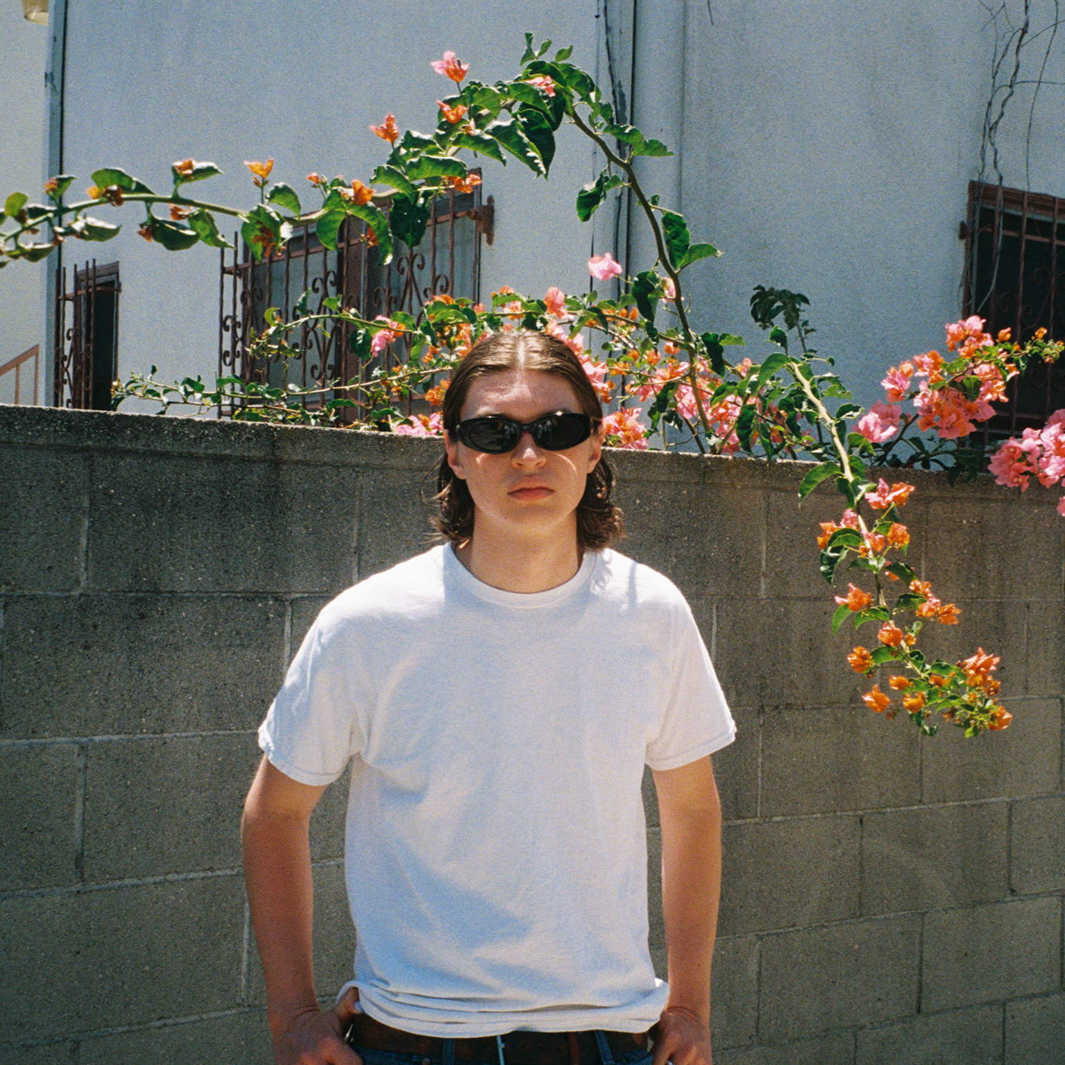 Person wearing sunglasses and a white t-shirt standing in front of a wall with flowering plants.