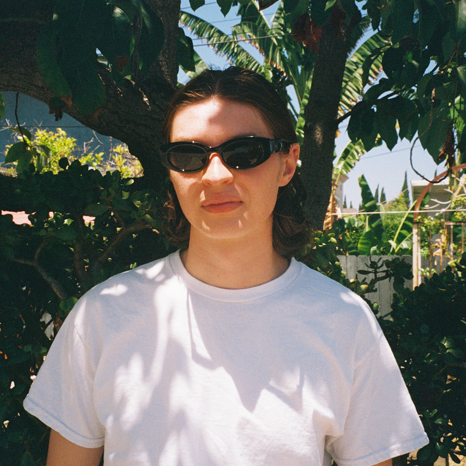 Person wearing sunglasses and a white t-shirt standing in front of green foliage