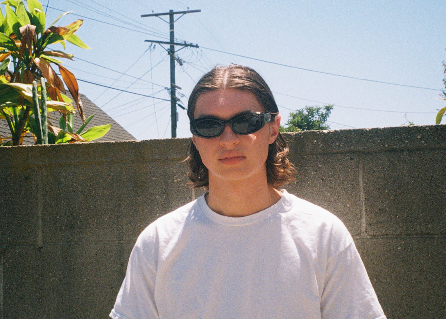 Person wearing sunglasses standing in front of a concrete wall with plants and power lines in the background