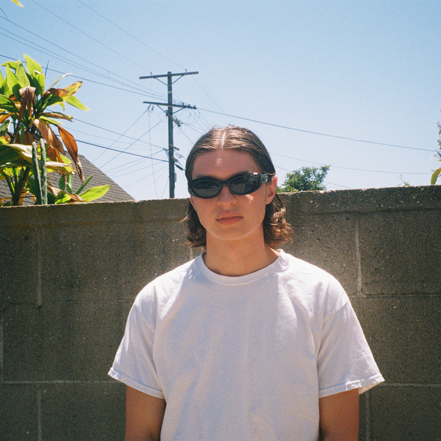 Person wearing sunglasses standing in front of a concrete wall with plants and power lines in the background