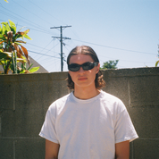 Person wearing sunglasses standing in front of a concrete wall with plants and power lines in the background