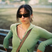 Woman in a green sweater with sunglasses and a brown strap over her shoulder, standing outdoors.