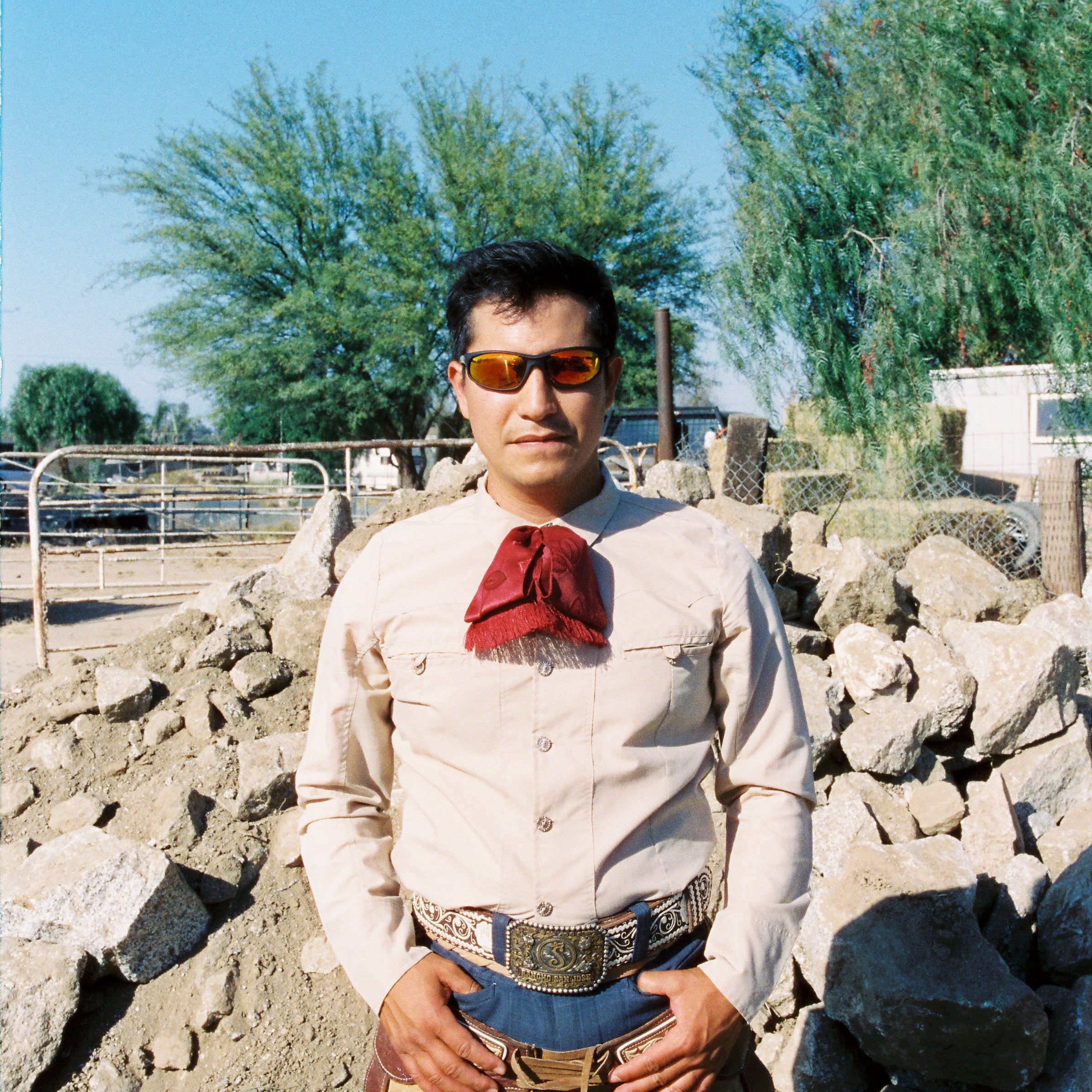 Man wearing a white shirt with a red handkerchief, sunglasses, and beige pants standing in front of rocks and trees.