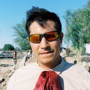 Man wearing sunglasses and a red scarf in an outdoor setting with trees and vehicles in the background.