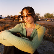 Person wearing sunglasses and a green sweater in an outdoor setting with vehicles and trees in the background.