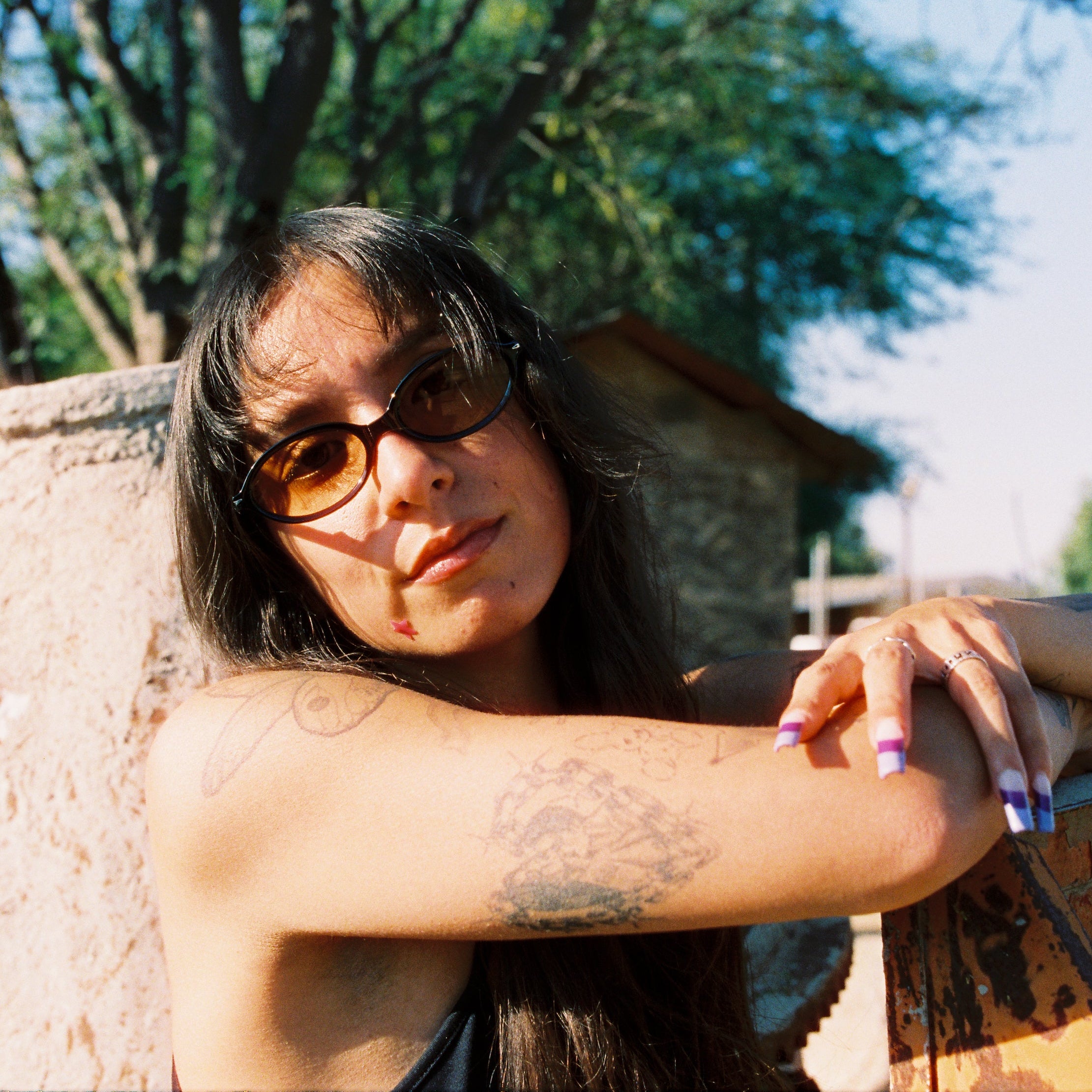 Person wearing sunglasses and a black dress with tattoos, sitting outdoors.